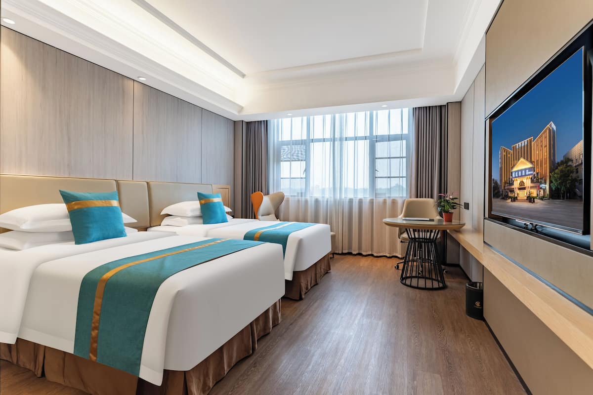 Ein Hotelzimmer mit zwei Betten, einem kleinen Tisch, einem Sessel, einem Fernseher und einem Fenster mit Vorhängen.