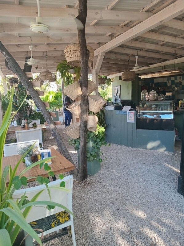 Café ao ar livre com mesas de madeira, cestos suspensos e balcão com mostrador de comida.