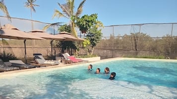 A swimming pool with lounge chairs and umbrellas, surrounded by palm trees and a fence.