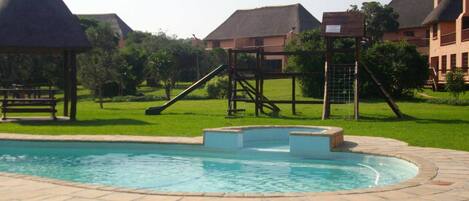 Una piscina con un deck perimetral, una estructura de juegos infantiles y edificios residenciales al fondo.