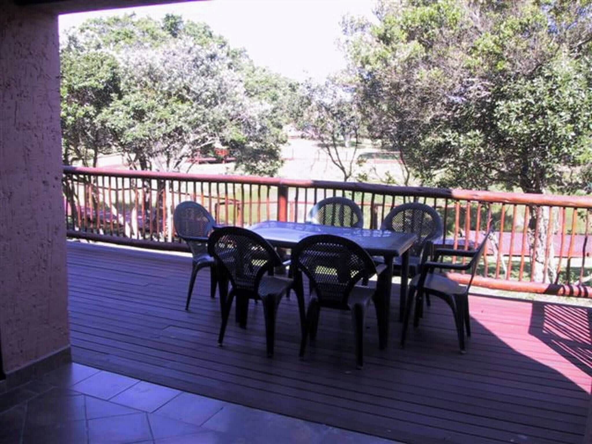 Un patio al aire libre con una mesa y sillas, rodeado por una baranda roja, con vista a un jardín con árboles y una cerca.