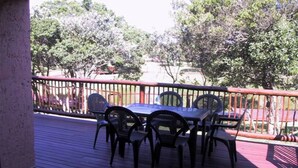 Un patio al aire libre con una mesa y sillas, rodeado por una baranda roja, con vista a un jardín con árboles y una cerca.
