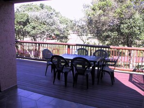 An outdoor patio with a table and chairs, surrounded by a red railing, overlooking a garden with trees and a fence.