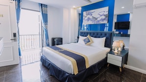 A hotel room with a large bed, a bedside table with a lamp, a flat-screen TV, and a window with curtains.