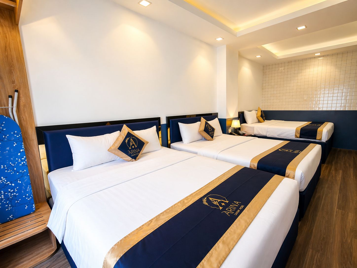 A hotel room with three beds, each with a navy blue headboard and white bedding. There is a wooden floor, a wall-mounted shelf, and a small table with a lamp.