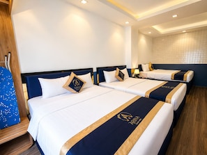 A hotel room with three beds, each with a navy blue headboard and white bedding. There is a wooden floor, a wall-mounted shelf, and a small table with a lamp. - Arina Hotel 3 (Tan Ninh)