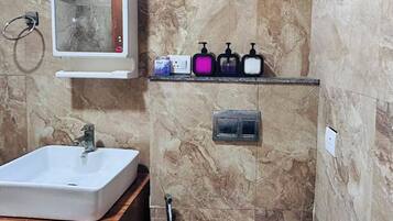 Un baño con lavamanos blanco, un espejo, un estante con dispensadores de jabón, ducha e inodoro.