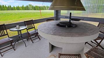 A covered outdoor seating area with a round table and chairs, a grill, and a view of a green field.