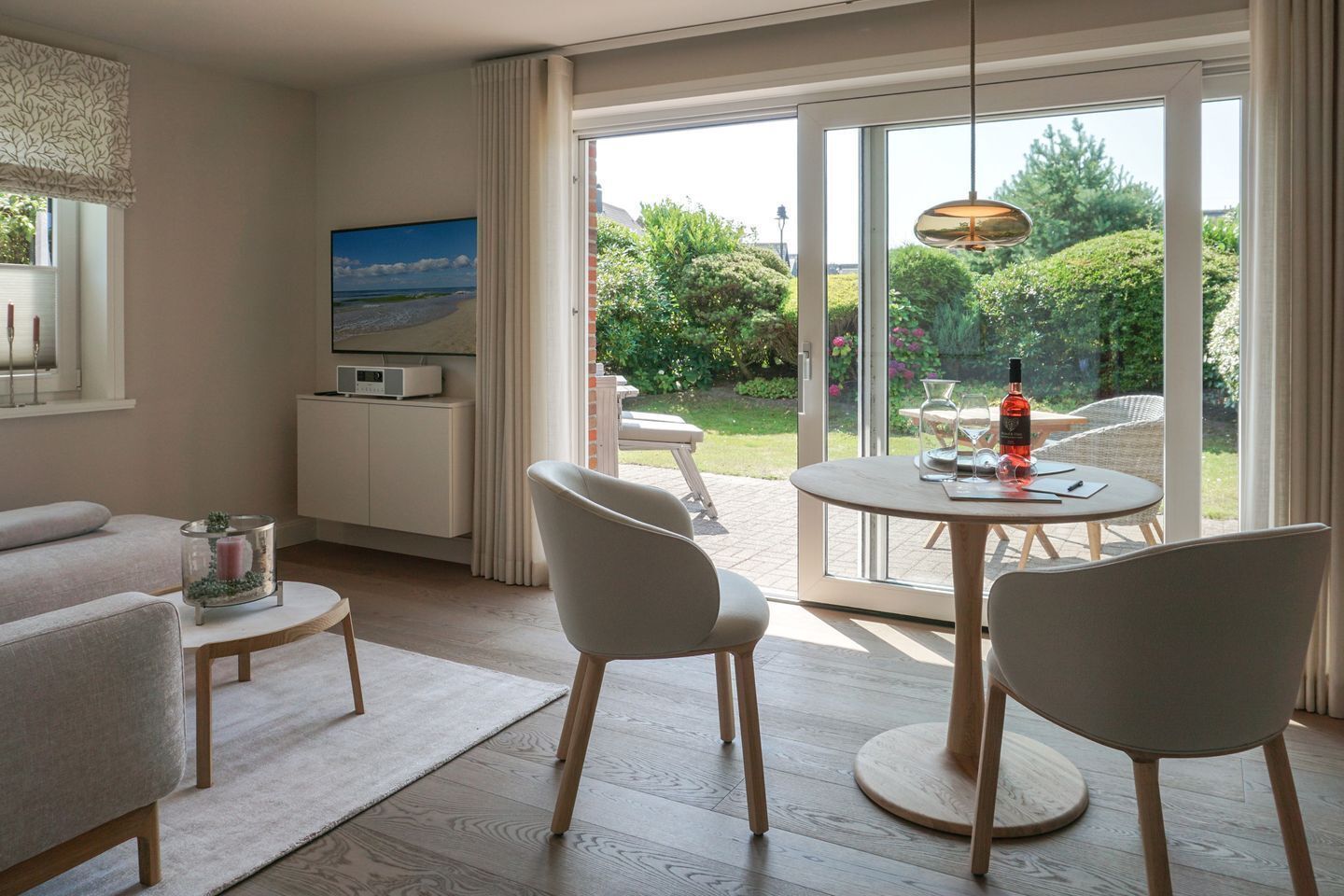 Een moderne woonkamer met een bank, een ronde tafel met stoelen, een salontafel met glazen blad, een televisie en uitzicht op een tuin door grote schuifdeuren.