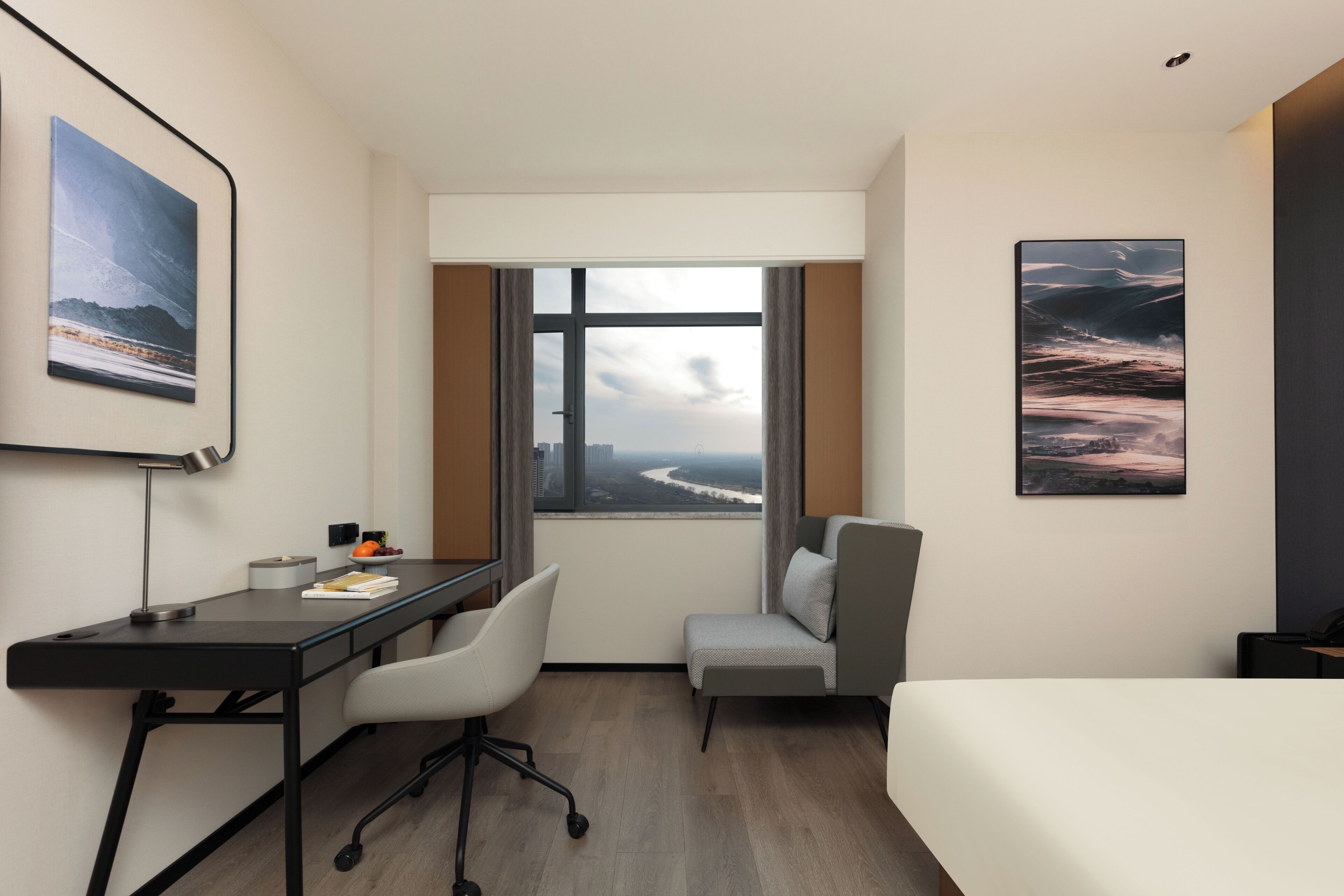Habitación de hotel con escritorio, silla, cama y ventanas amplias que ofrecen vistas a un paisaje urbano.
