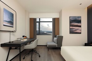A hotel room with a desk, chair, bed, and large windows offering a view of a cityscape.