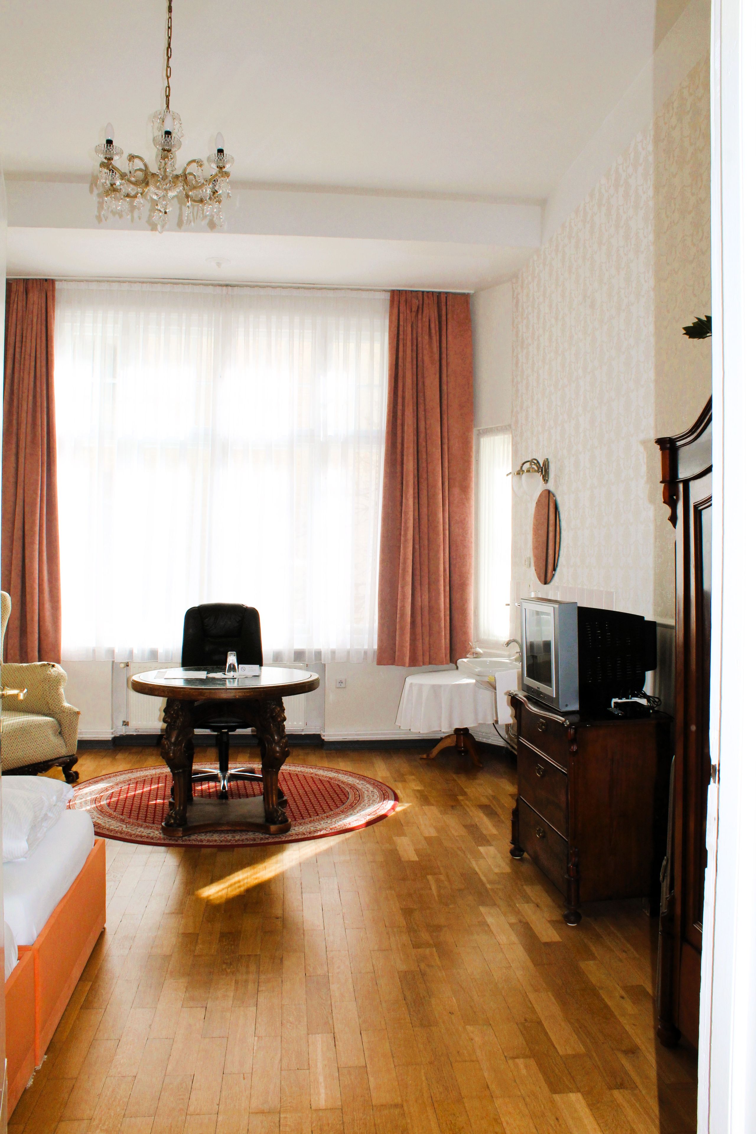 A hotel room with a wooden floor, a round table, a chair, a bed, a television, and a chandelier.