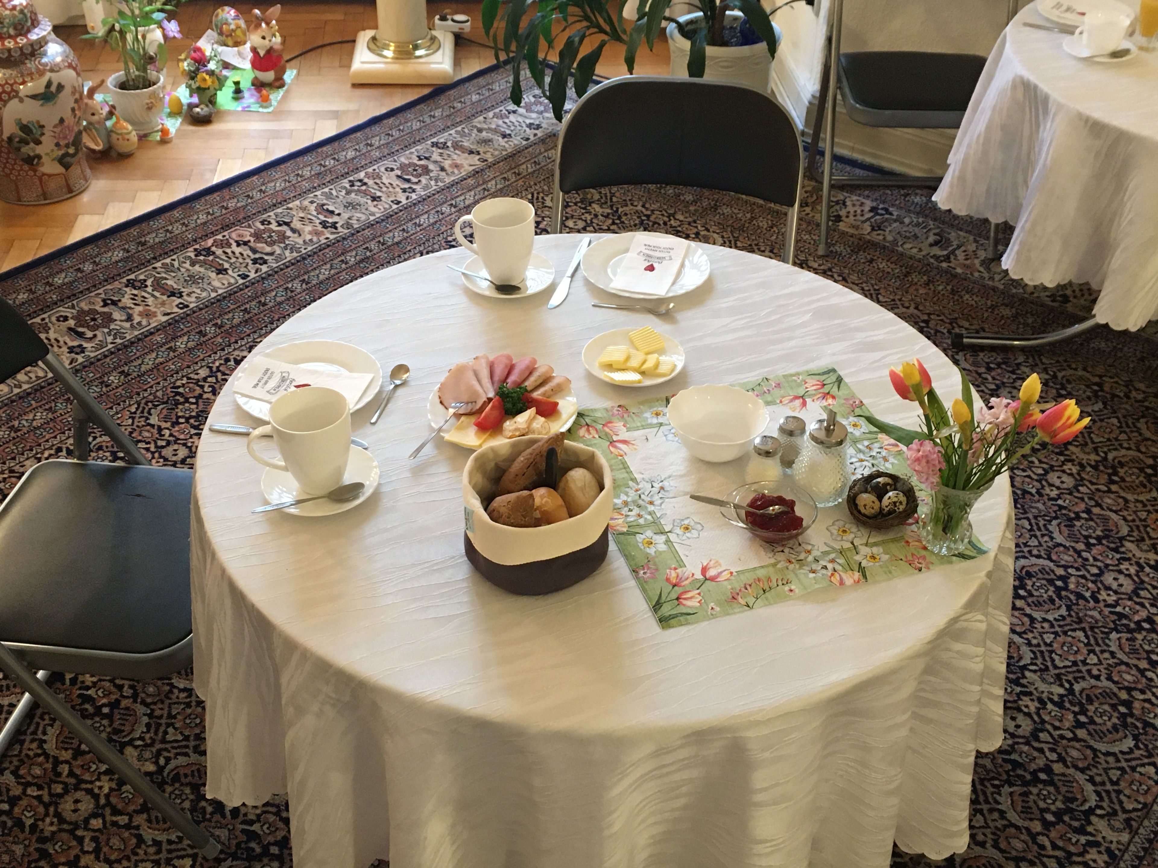 A round dining table with a floral tablecloth, a bowl of bread, a plate of sliced ham and cheese, and a vase with flowers.