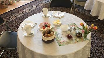 A round dining table with a floral tablecloth, a bowl of bread, a plate of sliced ham and cheese, and a vase with flowers.