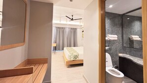Una camera d'albergo moderna con una panca in legno, un letto con copripiumino a motivi verdi, una ventola a soffitto e un bagno con un piano di lavoro in marmo e un water.