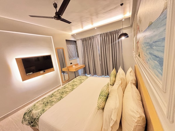 A hotel room with a bed, a TV mounted on the wall, a ceiling fan, a small table, and a painting on the wall.
