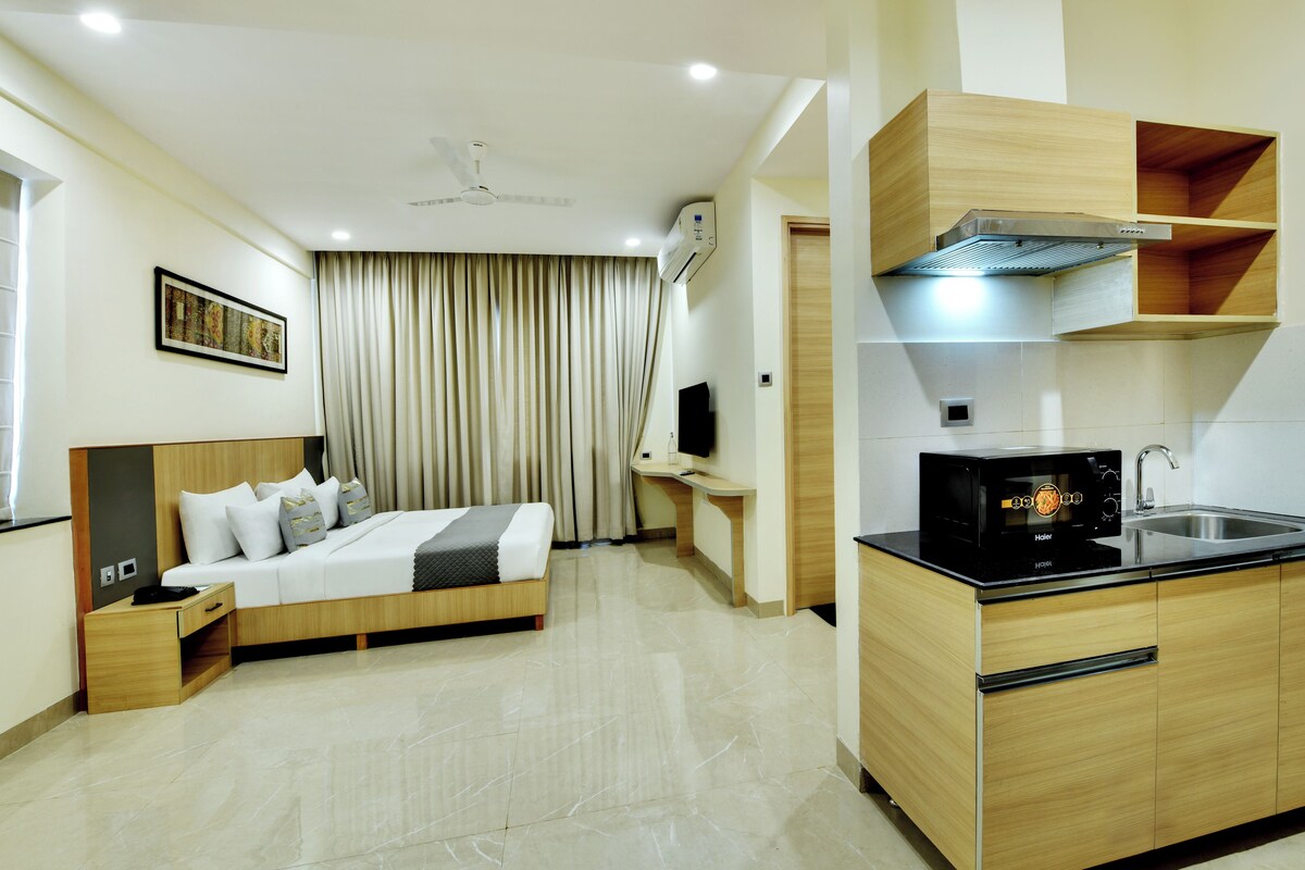 A hotel room with a bed, bedside table, a desk, a chair, a television, and a kitchenette with a microwave and sink.