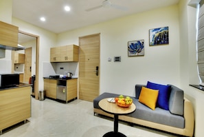 A modern living room with a grey sofa, a small round table with fruit, and a kitchen area with wooden cabinets and a black countertop. - Westside Studio & Apart Hotel (Hyderabad)