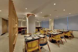 A modern dining area with multiple tables and chairs, a wooden partition, and a large window with blinds. - Westside Studio & Apart Hotel (Hyderabad)
