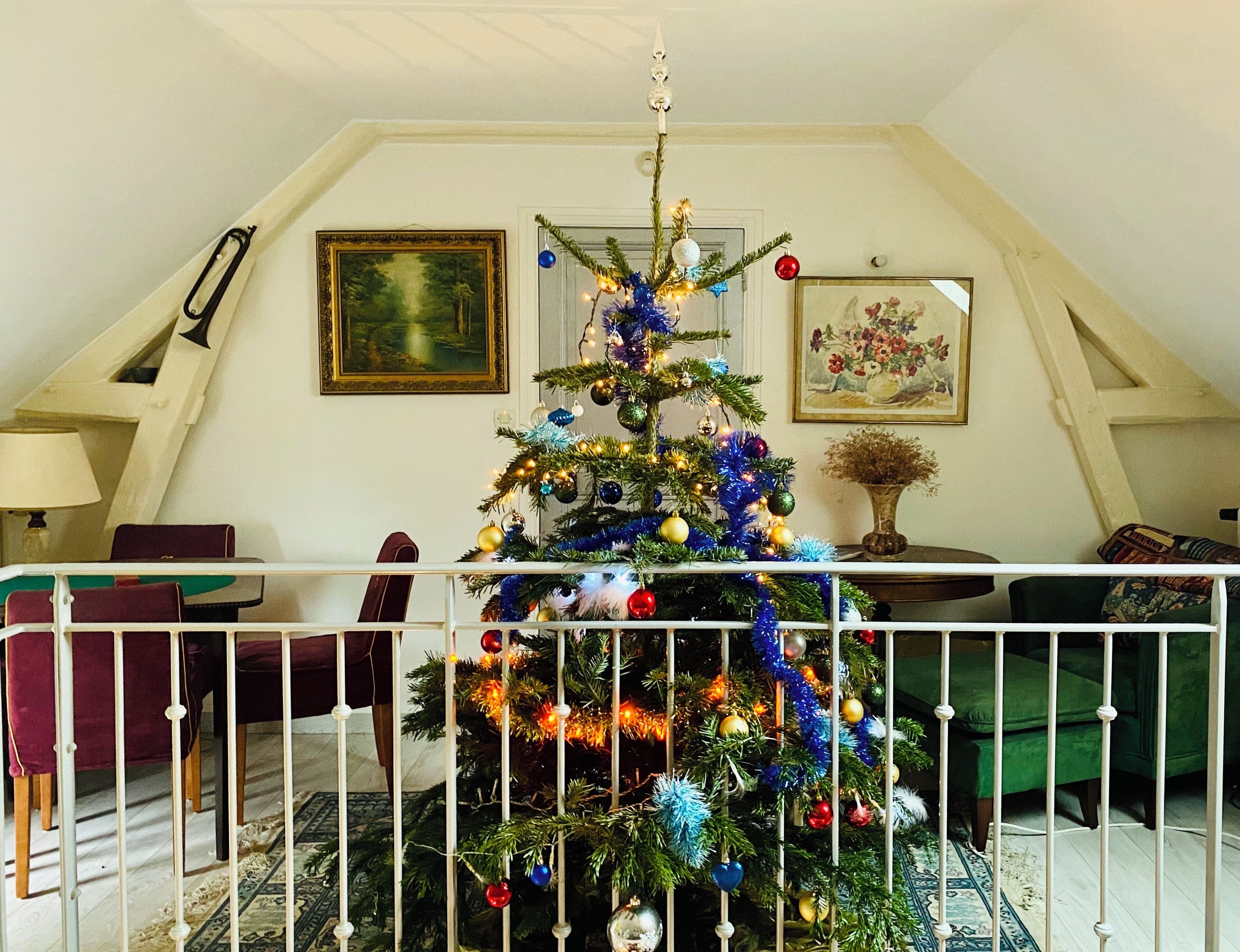 Un sapin de Noël décoré dans une pièce dont le plafond est en pente, avec un tableau encadré et un vase contenant des fleurs séchées.