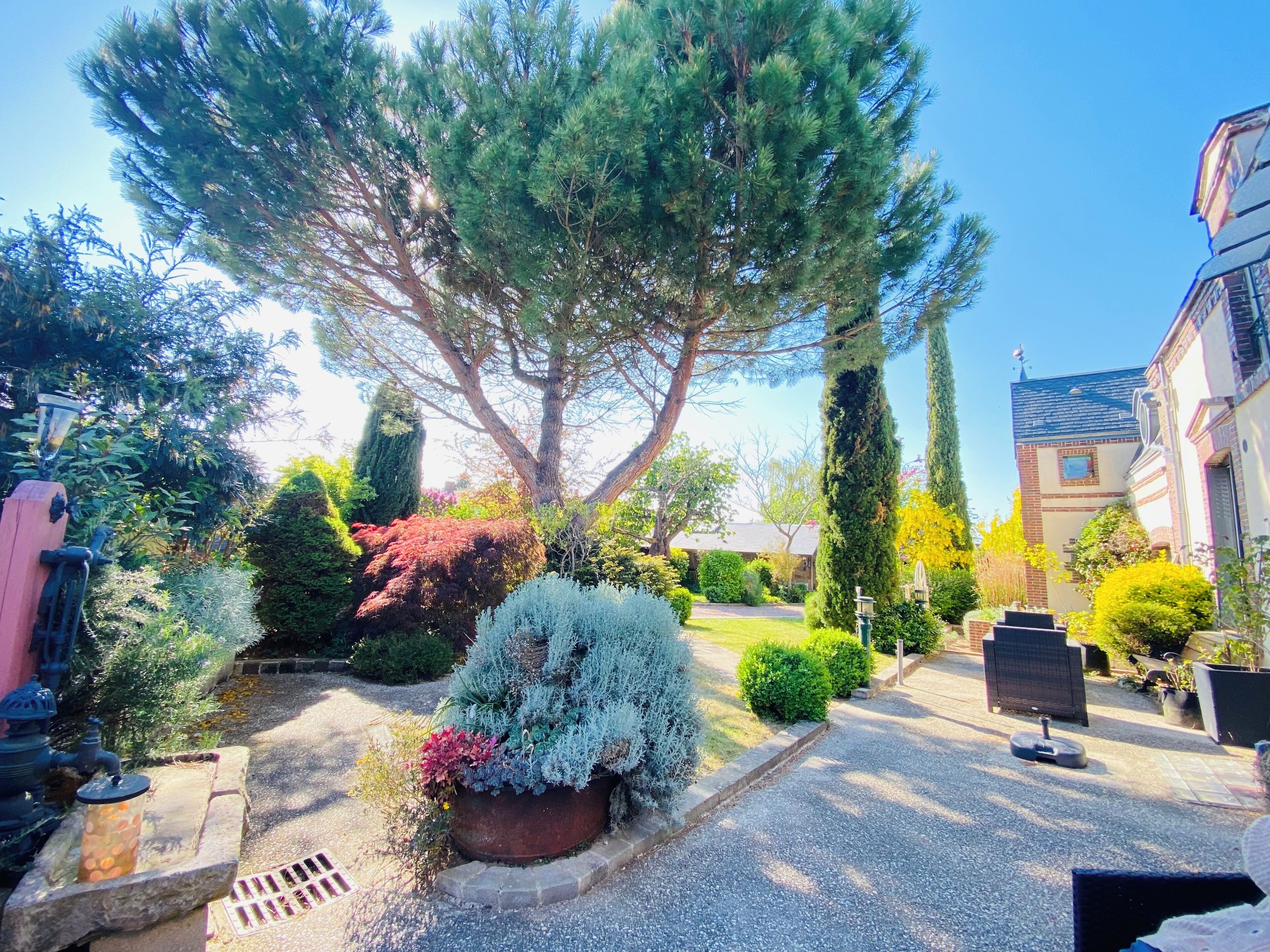 Un jardin résidentiel agrémenté d’un grand arbre, de divers arbustes, d’un puits en pierre et d’une allée pavée.