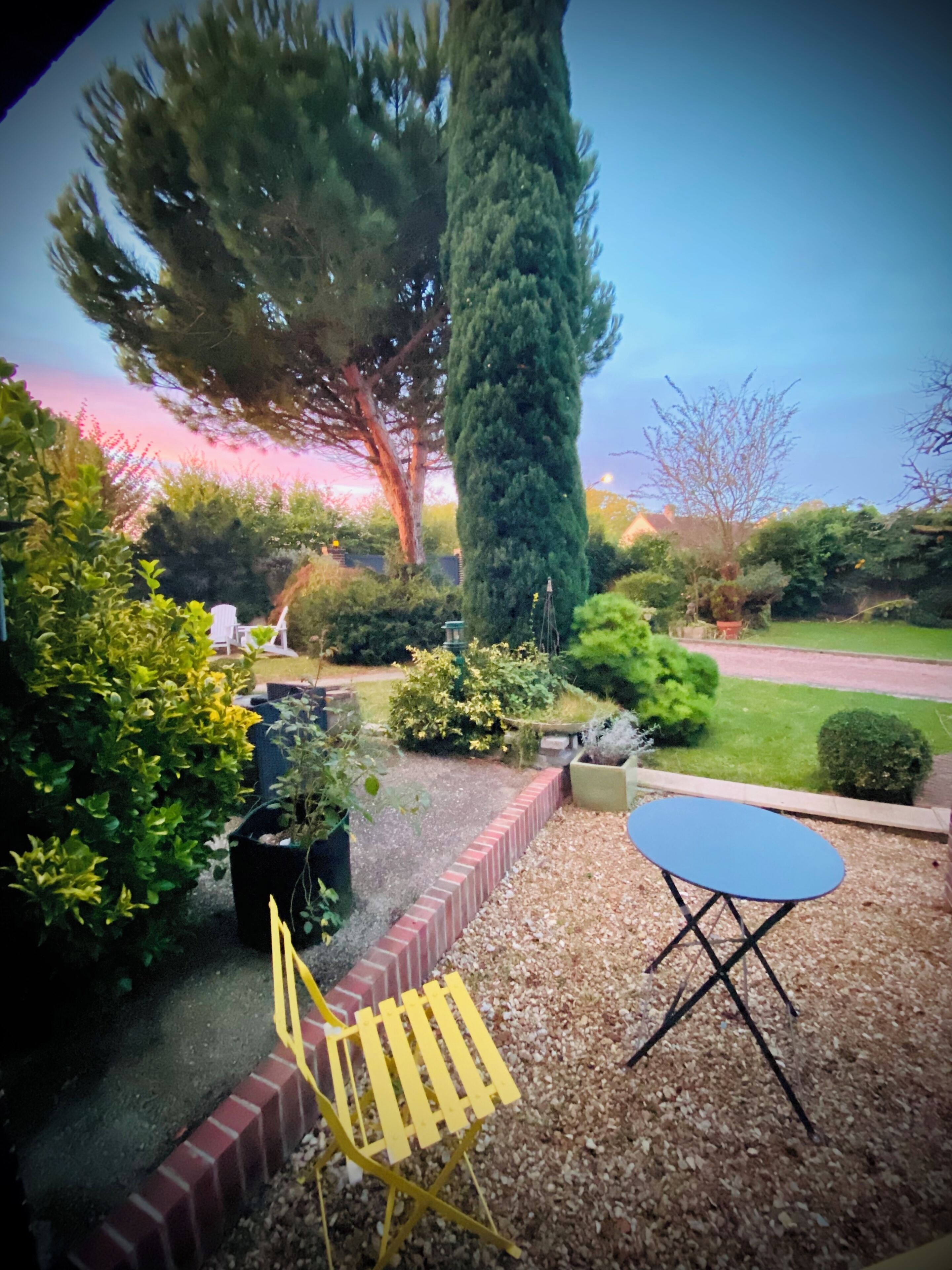 Un jardín con un sendero de ladrillos, una silla amarilla, una mesa azul y diversas plantas.
