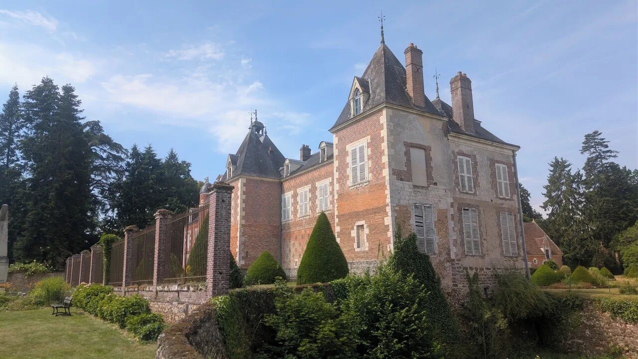 Un grand bâtiment historique, doté de nombreuses cheminées, d’une tour imposante et d’un jardin bien entretenu avec des haies taillées.