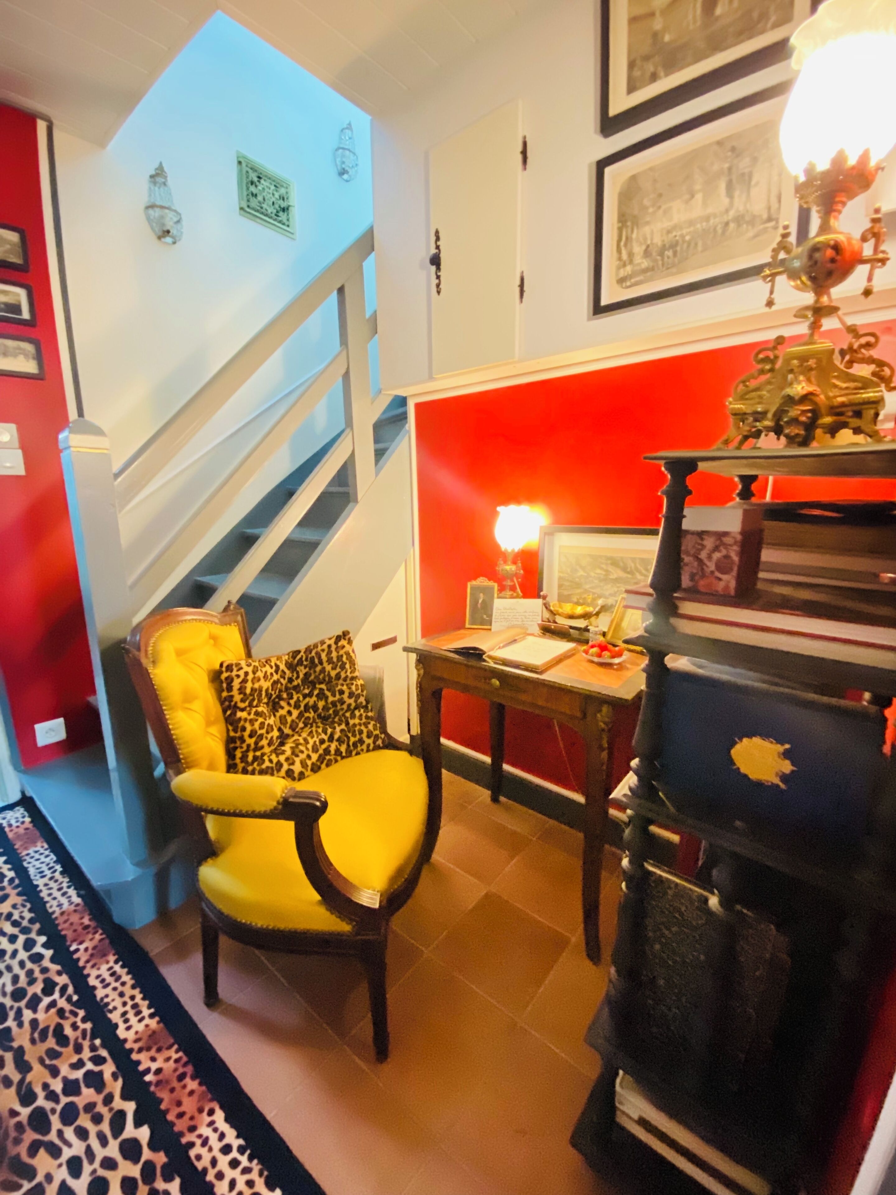 Una habitación con una escalera, un sillón amarillo, una mesa con una lámpara y una estantería.