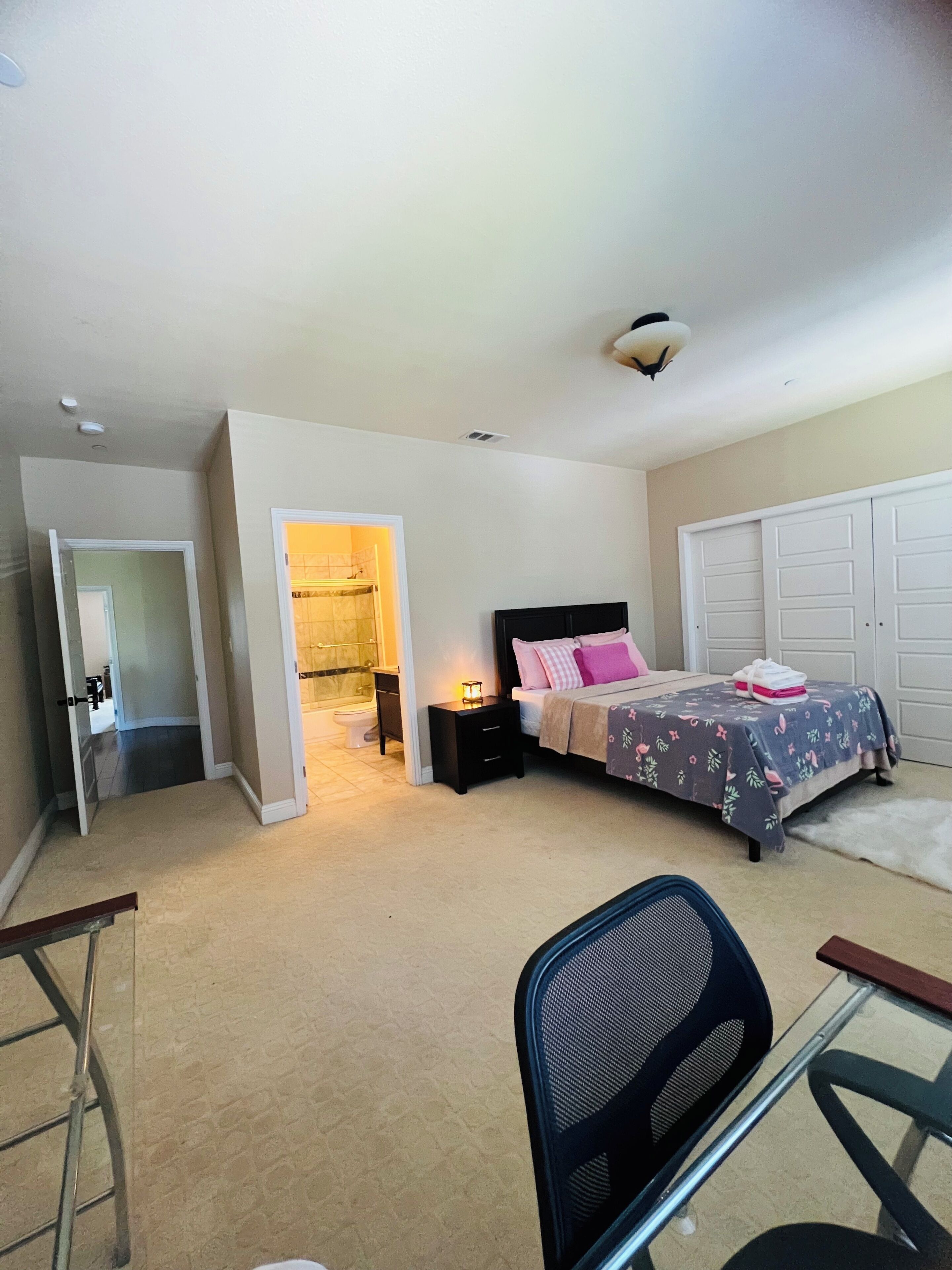 Une chambre à coucher avec un lit, des tables de chevet, un bureau et une chaise. On peut voir une armoire et une salle de bain à travers une porte ouverte.