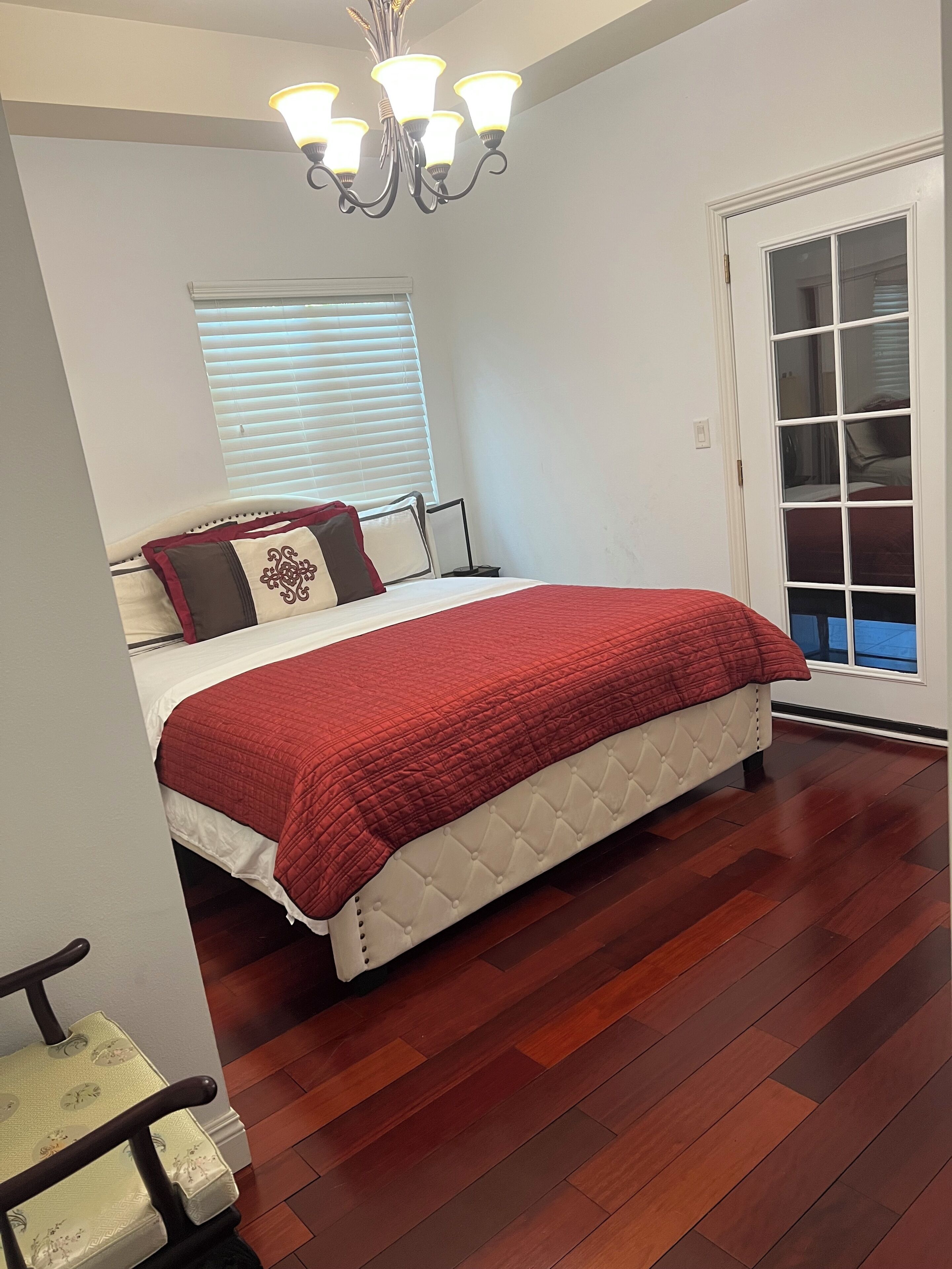 A bedroom with a bed, a chandelier, a window with blinds, and a door with a glass panel.