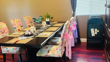 Un comedor con una mesa larga, sillas decoradas con cintas rosas, una planta pequeña y un mueble negro con papeles.