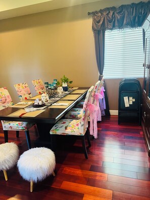 Un coin repas avec une longue table, des chaises ornées de rubans roses, une petite plante et un buffet noir contenant des papiers.