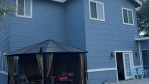 A two-story blue house with a covered patio area, a red grill, and a door leading to a garage.