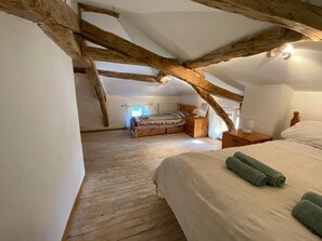 A bedroom with a sloped ceiling, wooden beams, a bed with white linens, and a window with curtains.