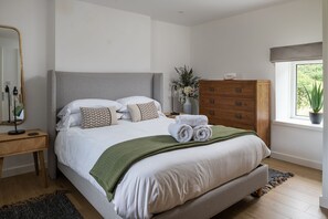 A bedroom with a bed, a dresser, a nightstand, a mirror, a window with a plant, and a vase with flowers. - Bayholme House - Luxury Cottages (Oxwich)