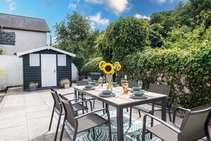 An outdoor patio with a table set for dining, surrounded by chairs, and a small shed in the background. - Bayholme House - Luxury Cottages (Oxwich)