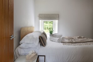 A bedroom with a bed, pillows, a window with a view, a bedside table, and a door. - Bayholme House - Luxury Cottages (Oxwich)