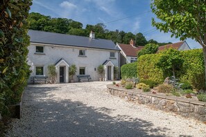 A two-story house with a gravel driveway, a stone wall, and a variety of plants and shrubs. - Bayholme House - Luxury Cottages (Oxwich)