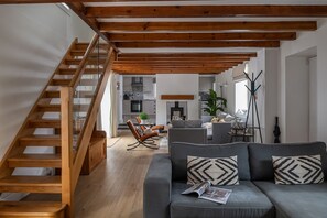 A modern living room with a wooden staircase, a grey sofa with patterned cushions, and a kitchen in the background. - Bayholme House - Luxury Cottages (Oxwich)