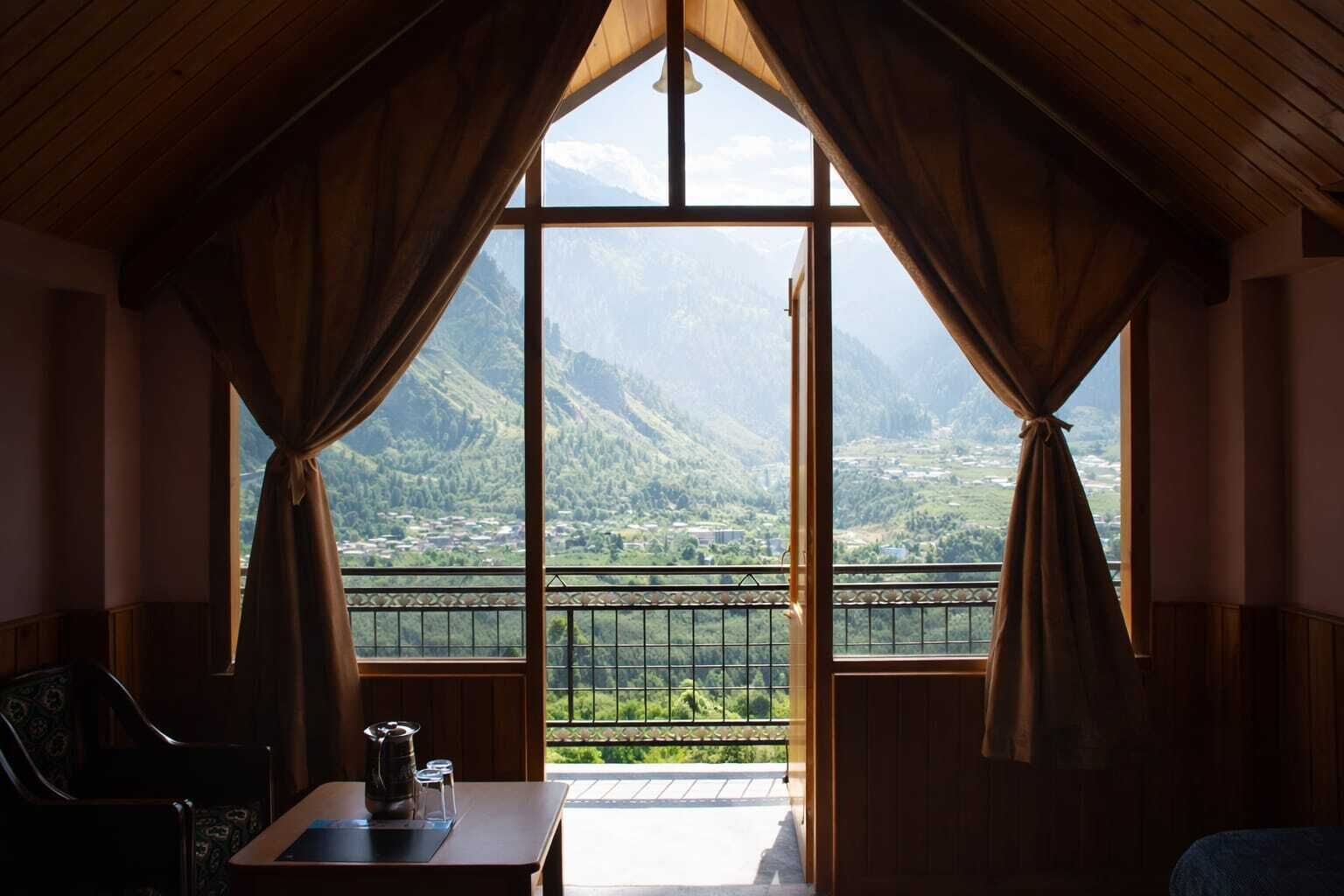 A room with a large window, curtains, a table with a teapot, and a chair.