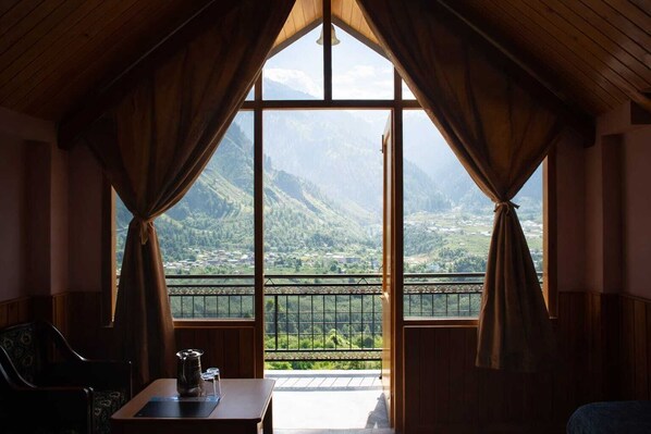 A room with a large window, curtains, a table with a teapot, and a chair.