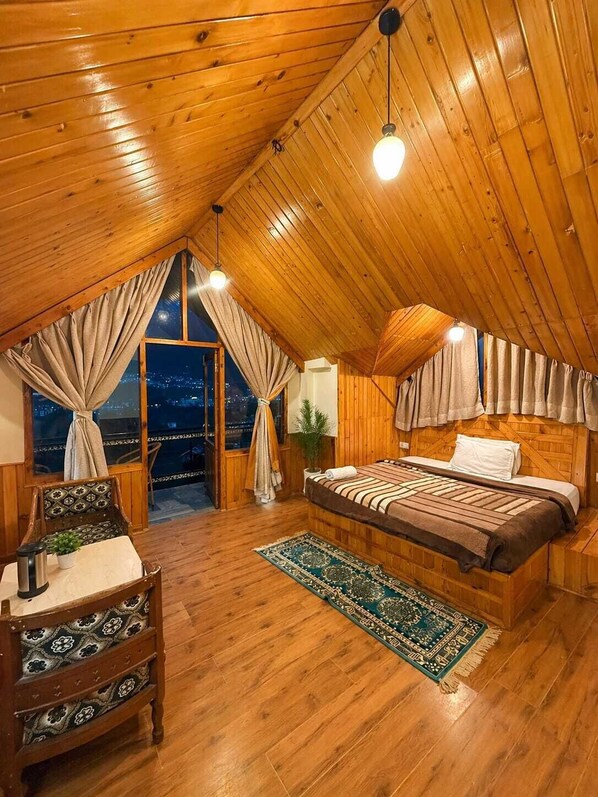 Ein gemütliches Schlafzimmer im Dachgeschoss mit Holzbalkendecke, einem großen Bett, einem Nachttisch, einem Sessel und Blick auf die beleuchtete Stadt.
