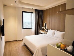 A hotel room with a bed, a bedside table with a mirror, a flat-screen TV mounted on the wall, and a window with a view of the city. - DOM HOTEL DALAT (Lam Vien - Da Lat)