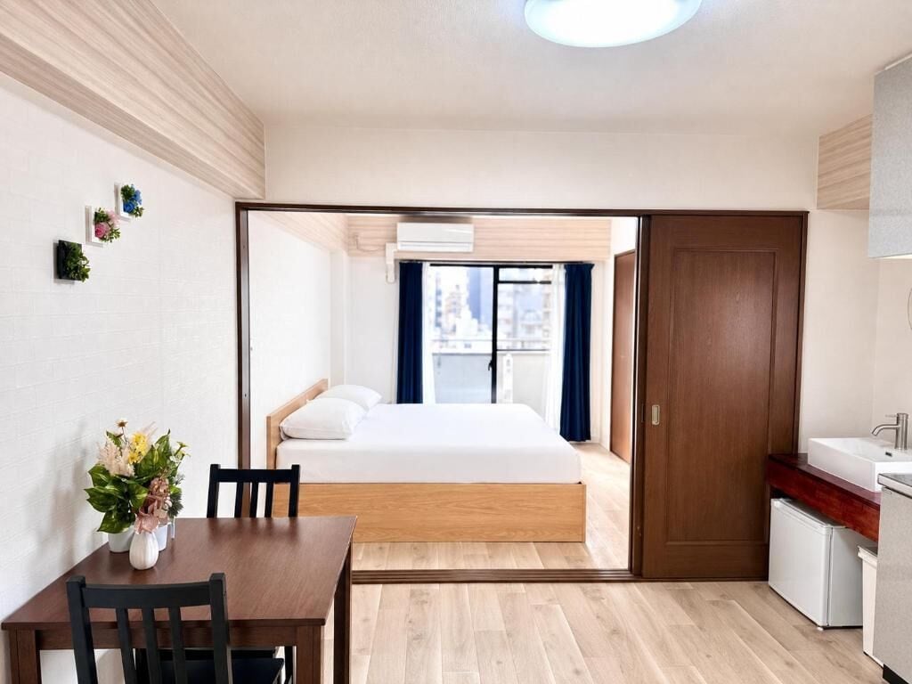 A modern hotel room with a wooden floor, a bed with white bedding, a dining table with chairs, a small kitchen area, and a view of the city through the window.