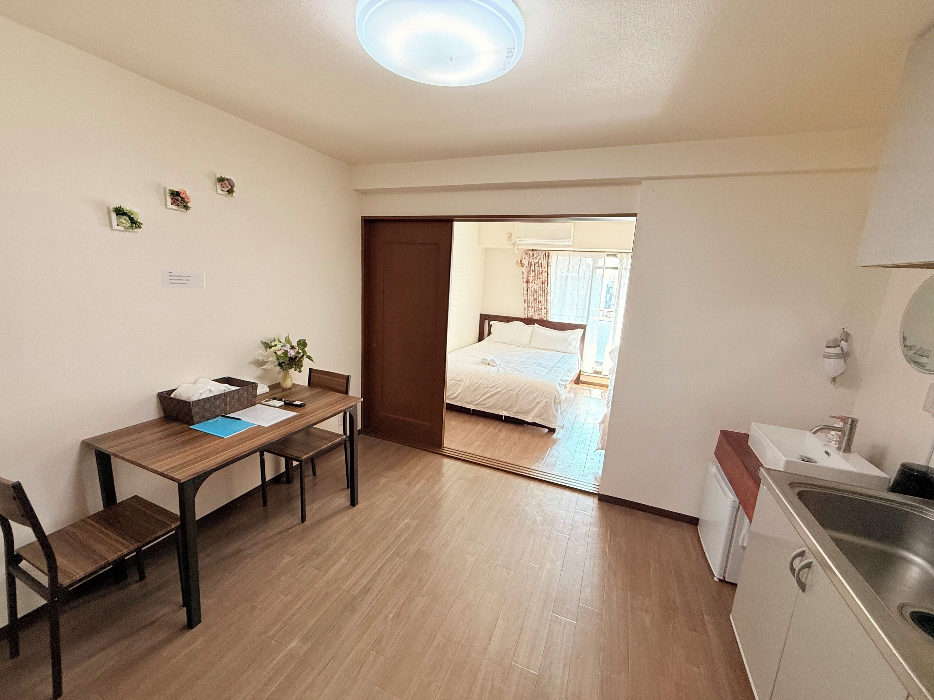 A compact room with a wooden dining table, a single chair, a small kitchen area with a sink, and a bed visible through an open door.