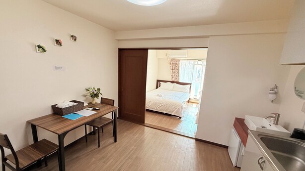 A compact room with a wooden dining table, a single chair, a small kitchen area with a sink, and a bed visible through an open door. - Raku Osaka Daikokucho Namba Shinsaibashi (Osaka)