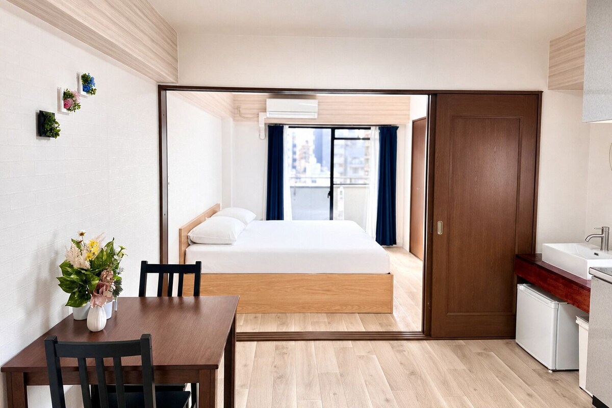 A hotel room with a bed, a dining table, a kitchen area, and a view of the city through the window.