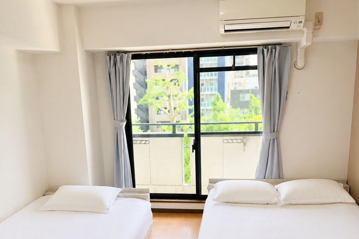 A hotel room with two beds, a window with curtains, an air conditioning unit, and a view of buildings outside.