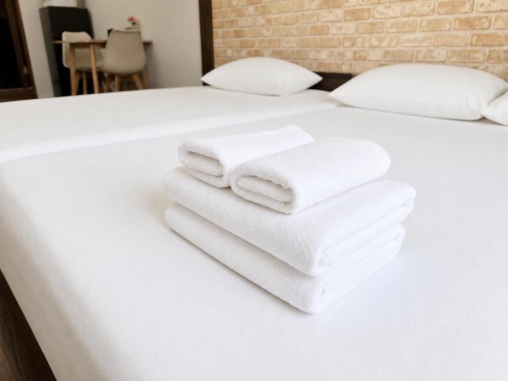 Een net opgemaakt bed met witte lakens en kussens, een bakstenen muur op de achtergrond, en een klein tafeltje met stoelen in de kamer.