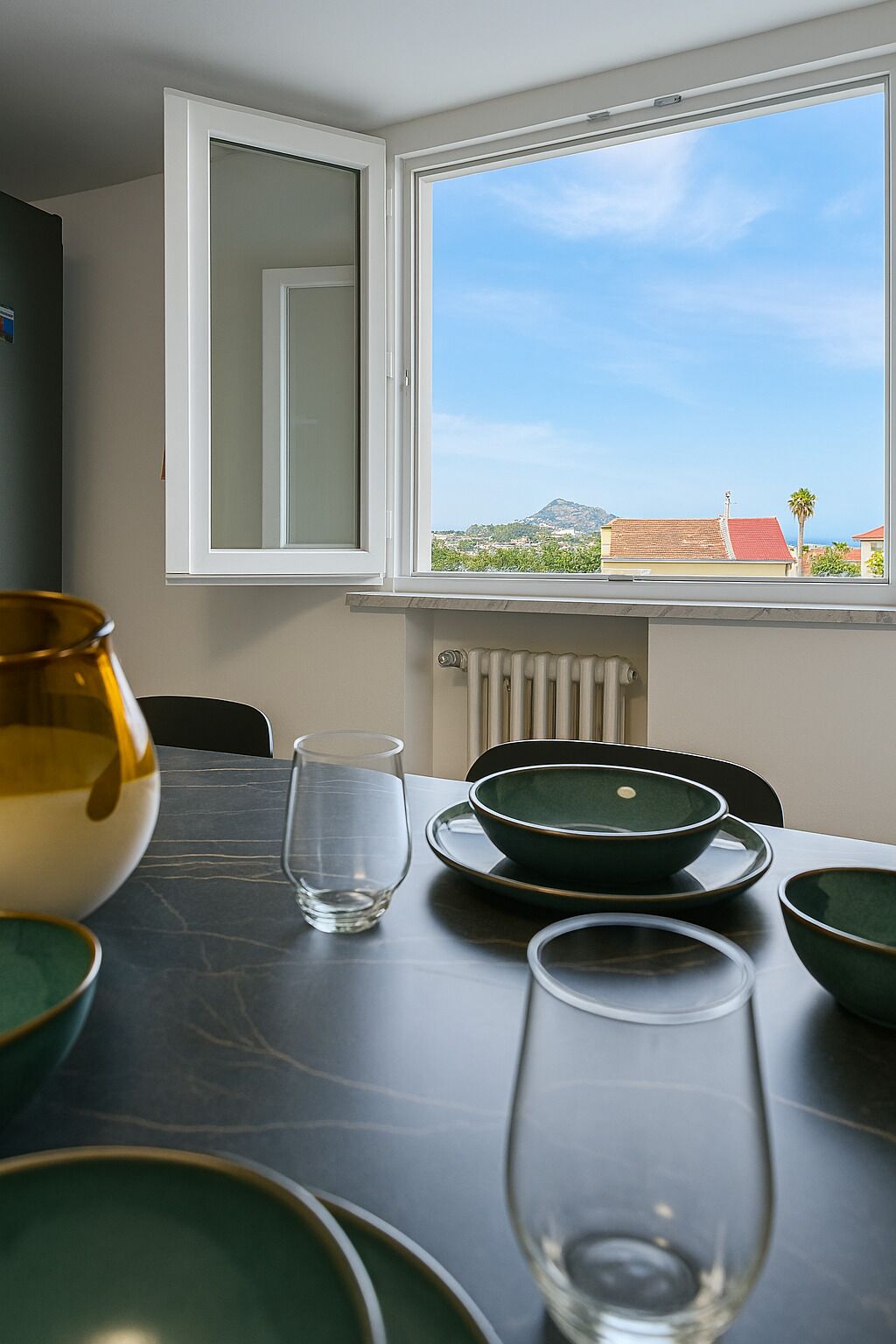 Ein Essbereich mit einem Tisch für zwei Personen, einem Fenster mit Blick auf Gebäude und einen Berg sowie einem Heizkörper unter dem Fenster.